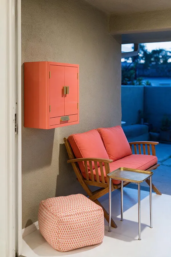 A compact eclectic patio features a coral wall cabinet mounted on a textured stucco wall, a wooden slatted loveseat with coral cushions, a small brass-topped side table, and a patterned pink ottoman on a pale floor. The space opens to a blue-toned outdoor view, creating a colorful, relaxed sitting area.