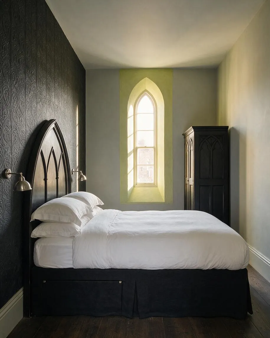 A narrow guest bedroom with a black arched bed frame against a textured dark accent wall, dressed in crisp white bedding. A tall pointed-arch window brings in soft daylight, and a carved black wardrobe adds to the Gothic feel.