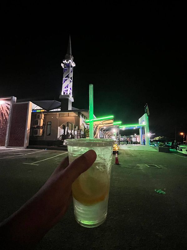 A hand holds a cup of lemonade in front of a brightly lit building at night.