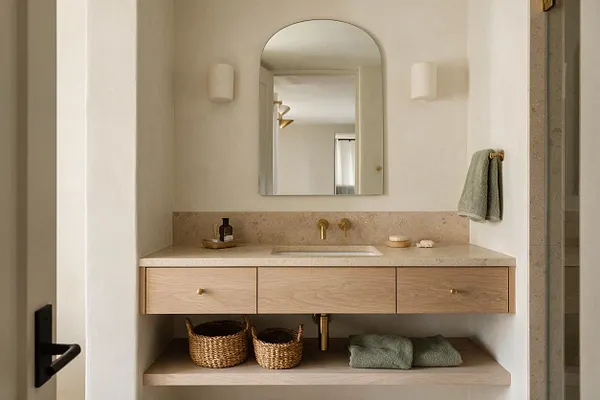 A serene master bathroom vanity with a pale stone countertop, light wood floating cabinet, and arched mirror centered above a simple sink. Brass faucet hardware, woven baskets, and soft green towels add warm organic texture against creamy walls.