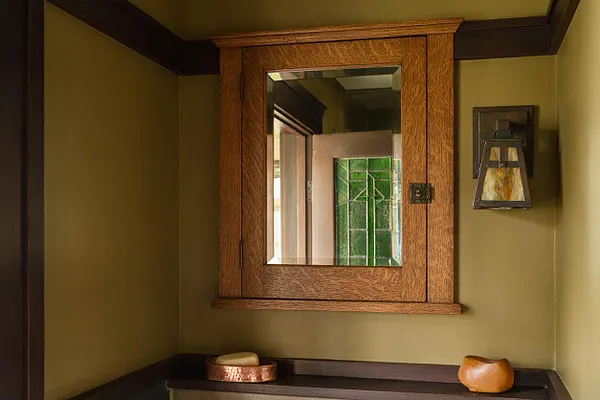 A Craftsman-style bathroom wall with olive green paint, dark wood trim, and a centered wooden medicine cabinet with a mirror. A small wall sconce sits to the right, and the shelf below holds simple decorative bowls, creating a warm, earthy look.