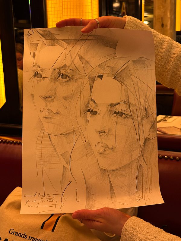 A hand holds a sketch of two stylized portraits in a warm, indoor setting.