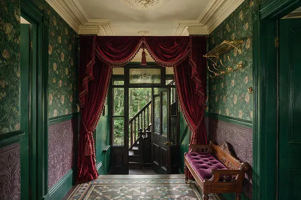 A narrow Victorian-style entryway with dark green patterned wallpaper, mauve embossed wall panels, and a patterned mosaic tile floor. Deep burgundy velvet drapes frame the doorway, and a carved wooden bench with a purple tufted cushion sits along one wall beneath a brass coat rack.