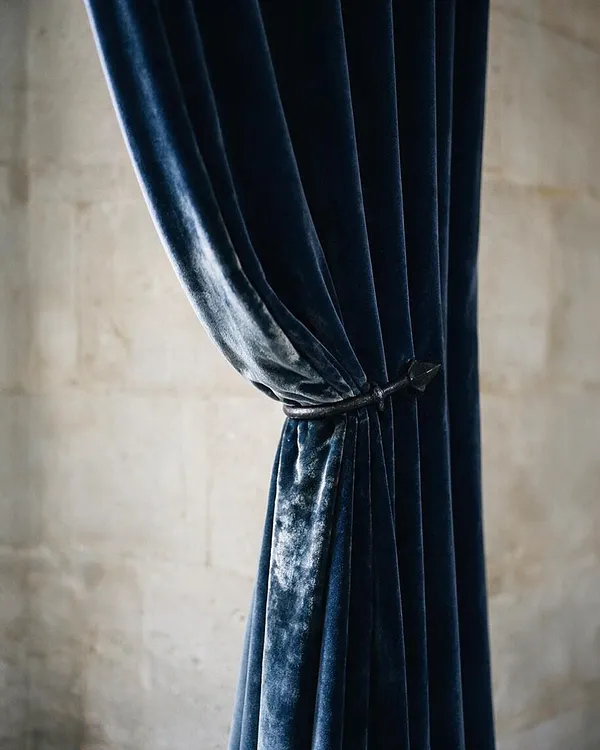 A close-up view of heavy dark blue velvet curtains gathered at the center with a small tieback. The plush fabric catches the light with a soft sheen, set against a muted stone or plaster wall in a moody Gothic-inspired interior.
