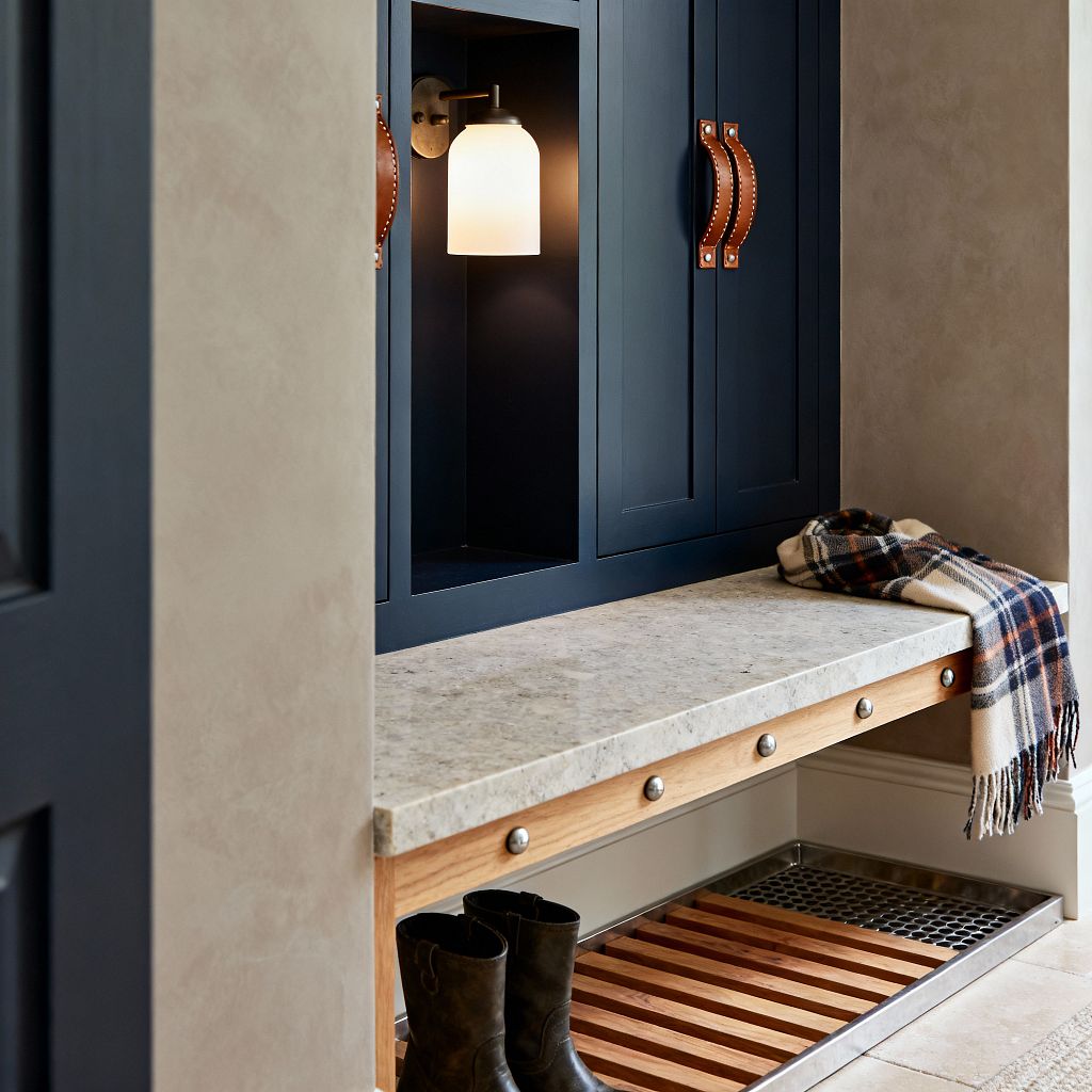 Close-up of a transitional mudroom built-in with deep navy Shaker cabinets and cognac leather strap handles. A light, speckled stone bench spans the unit, with an opal-glass brass sconce glowing inside a recessed cubby; below, a natural wood slatted shelf sits above a stainless boot tray, while warm beige walls and stone-look floor tiles complete the scene, with a plaid scarf on the bench and boots tucked underneath.