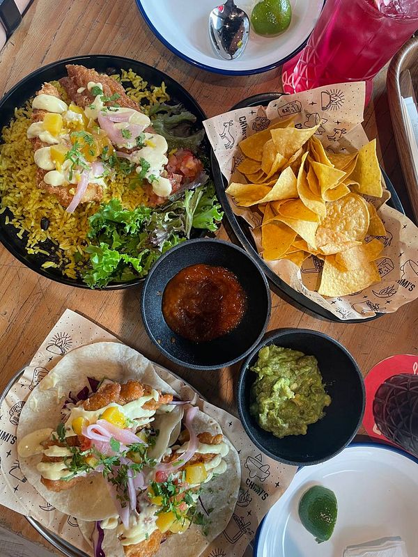 A wooden table set with Mexican lunch dishes including tacos, guacamole, salsa, and drinks is ready for a casual meeting or catch-up.