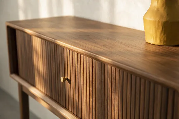 A close-up of a mid-century modern wooden console table with a smooth walnut top, vertically fluted cabinet doors, and a small brass knob. Warm sunlight casts soft shadows across the wood, and a yellow ceramic vase sits at the right edge.