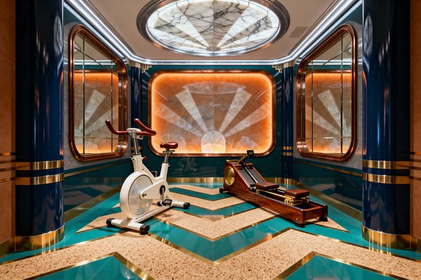 A luxurious Art Deco home gym with deep teal lacquered walls and brass-banded columns, a chevron floor combining teal panels, cork, and brass inlays, and a glowing amber onyx sunburst feature on the far wall. Overhead, a round marble light with radial segments crowns the ceiling. A white stationary bike with tan leather accents and a walnut-and-brass rowing machine sit centered, flanked by copper-framed mirrors and perimeter LED lighting.