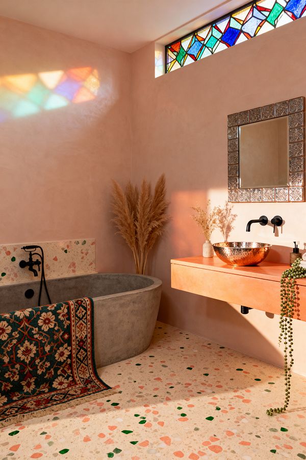 A bohemian bathroom with blush polished plaster walls and a cream terrazzo floor flecked with coral and green. A gray concrete soaking tub with matte black fixtures sits beside a peach floating vanity topped with a hammered copper vessel sink, an embossed metal-framed mirror, and dried grasses; a trailing succulent softens the edge. A patterned rug drapes over the tub, while a stained-glass clerestory window casts colorful light across the room.