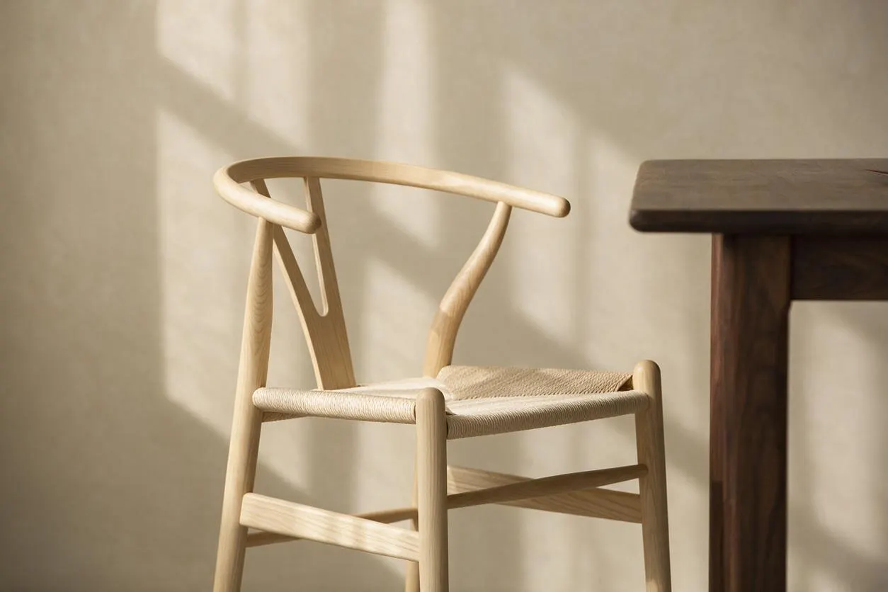 A light wood wishbone-style chair with a woven natural seat sits beside a dark wood table in warm sunlight. The neutral wall behind it casts soft shadows, emphasizing the chair’s sculptural frame and minimalist Japandi aesthetic.
