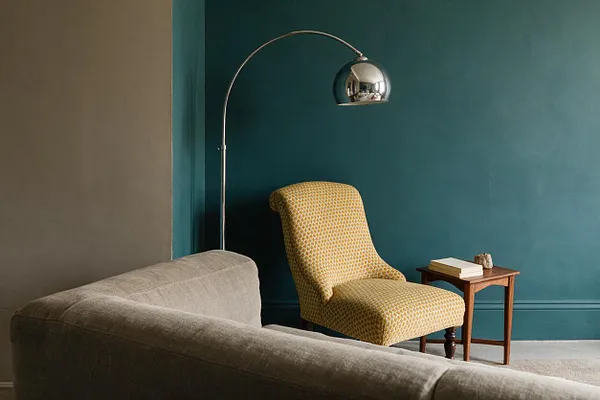 An eclectic living room vignette with a deep teal accent wall, a mustard patterned accent chair, and a chrome arched floor lamp. A neutral upholstered sofa fills the foreground, while a small wooden side table with books adds warmth beside the chair.