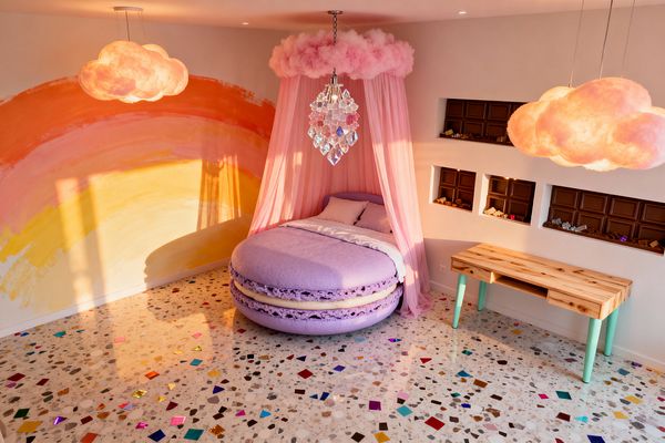 A whimsical kids' bedroom features a round lavender macaron-shaped bed wrapped in sheer pink canopy drapes beneath a cotton-candy cloud crown and crystal pendant. A hand-painted rainbow in peach, pink, and yellow spans one wall, while glowing cloud pendants, chocolate-bar recessed shelves filled with treats, a natural wood desk with mint legs, and a terrazzo floor flecked with multicolored confetti complete the playful Candy Land theme.
