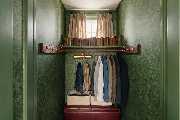 A narrow closet painted in deep green with patterned wallpaper and brass hardware, featuring a shelf lined with old books above a hanging rod. Light-colored curtains filter daylight from a small window, while neatly arranged shirts and jackets hang below, with storage bins and a burgundy leather bench at the bottom.