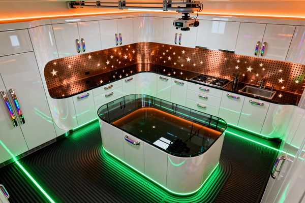 A futuristic kitchen with glossy white curved cabinets and iridescent handles, a perforated copper backsplash with star cutouts, and neon green LED lighting along the plinths and floor grooves. A central island has a smoked glass top with amber illumination inside, set over ribbed black flooring, with a stainless-steel sink, gas cooktop, and a ceiling-mounted device enhancing the high-tech atmosphere.