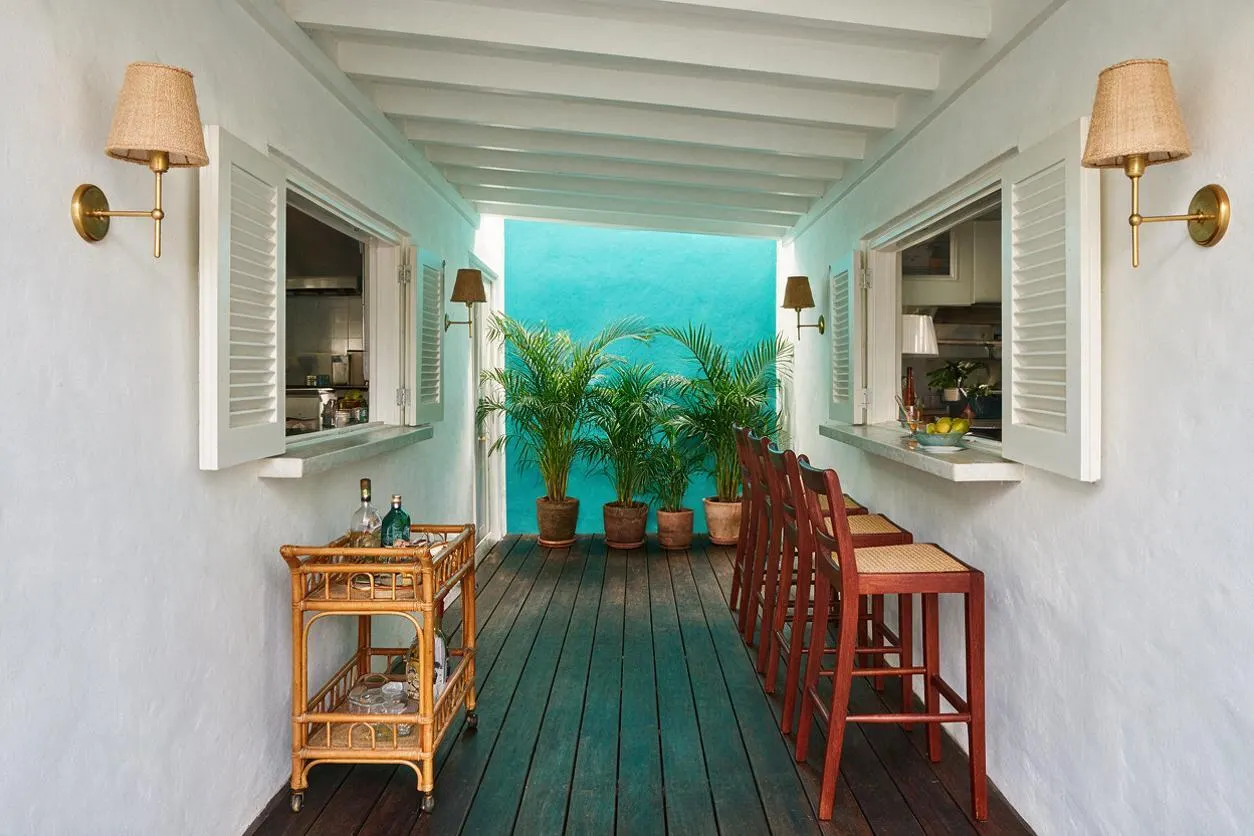 A narrow tropical backyard corridor with white stucco walls, open shuttered windows, and a turquoise end wall. Dark painted wood decking leads to potted palm plants, with a wicker bar cart on one side and tall red wooden stools on the other, creating a relaxed outdoor entertaining space.