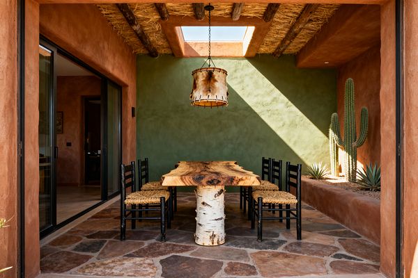 A Southwestern dining courtyard features a live-edge wooden table on a birch log base beneath a rawhide drum pendant, set before a sage green stucco wall with a raised planter of cacti and agave. Terracotta plaster walls, exposed timber-and-thatch rafters with a rectangular skylight, black ladder-back chairs with woven seats, and variegated flagstone flooring complete the sunlit space accessed by large sliding glass doors.