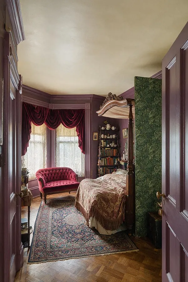 A compact Victorian-style studio apartment with mauve painted trim and walls, a tufted burgundy loveseat by lace-draped windows, and a carved canopy bed with patterned bedding. A floral green partition, antique bookshelf, oriental rug, and parquet wood floor create a richly layered, old-world atmosphere.