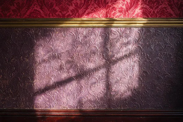 A close-up of a Victorian-style wall treatment featuring embossed damask wallpaper in mauve and plum tones, framed by a gold trim near the top and dark wooden wainscoting below. Soft light and shadow fall across the textured surface, highlighting the ornate floral pattern and rich, moody color palette.