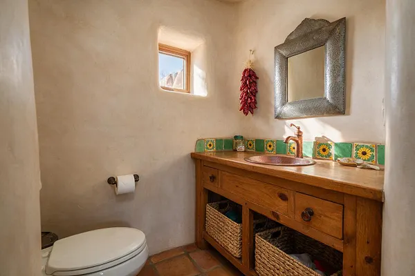 A small Southwestern-style powder room with plaster walls, a warm wood vanity, copper sink and faucet, and a green Talavera tile backsplash with sunflower motifs. The room includes a silver-toned framed mirror, a tiny window admitting daylight, terracotta floor tiles, woven storage baskets, and hanging red dried chiles for a rustic accent.
