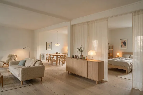 A bright Scandinavian studio apartment with pale wood floors, white walls, and soft beige textiles. A living area with a textured sofa sits beside a dining nook, while sheer curtains separate the bedroom with a low wooden bed and warm bedside lighting.