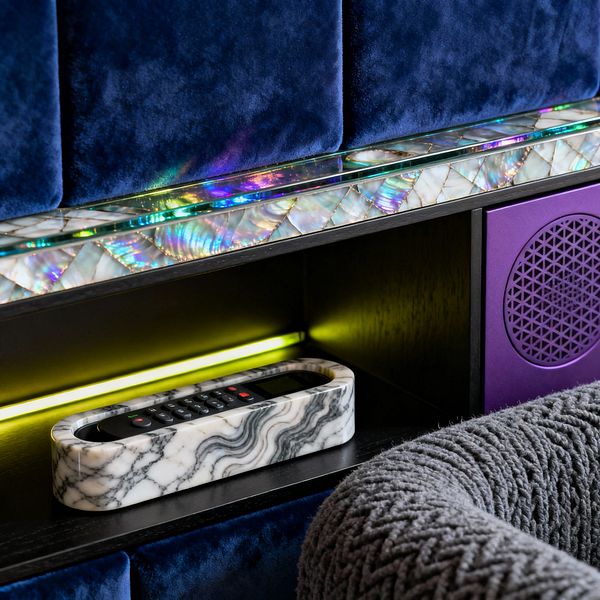 Close-up of a psychedelic media room wall with deep navy velvet panels above a band of quilted mother-of-pearl topped by a prismatic glass ledge. A black-stained shelf lit by chartreuse LED holds a gray-and-white marble tray containing a remote. To the right, a matte purple speaker with a geometric perforated grille sits beside a chunky gray knit armrest, creating a layered mix of color, light, and texture.