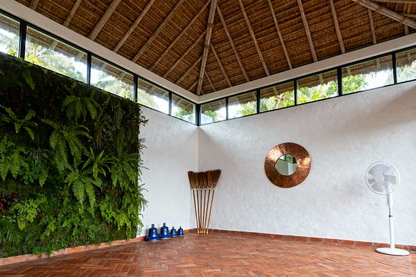 A tropical home gym with a tall living plant wall of ferns on the left, herringbone terracotta tile floor, and white textured plaster walls beneath a steep thatched ceiling with clerestory windows. In the corner sit four blue kettlebells and a bundle of straw brooms; a hammered copper round mirror hangs on the wall, and a white pedestal fan stands to the right, signaling natural airflow and an open, breezy atmosphere.
