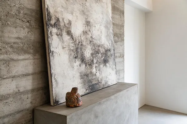 A minimalist Brutalist interior vignette with a textured concrete wall and matching concrete pedestal. A large abstract monochrome painting leans against the wall, and a small rustic ceramic vessel sits in front of it. Soft natural light enters from the right, highlighting the raw surfaces and neutral palette.