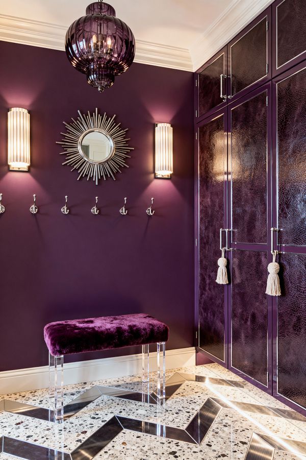 A mudroom painted deep plum features glossy purple built-in cabinets with textured leather-like panels, chrome pulls, and cream tassel ties. Two vertical ribbed-glass sconces flank a silver sunburst mirror above a row of polished chrome wall hooks. An amethyst glass pendant hangs from a white ceiling with crown molding. A plush purple bench with clear lucite legs stands on speckled terrazzo flooring with stainless chevron inlays, softly lit and reflective.