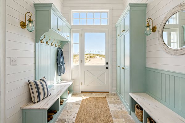 A bright coastal mudroom with white shiplap walls features seafoam green built-in lockers and benches flanking a white door with a large window and transom. Brass hooks, small brass knobs, and aqua seeded-glass sconces add warm sparkle. The benches have whitewashed wood tops and cubbies with woven baskets and shoes; a striped pillow and a denim jacket rest casually. A tumbled stone floor and jute runner lead to a view of sand dunes outside.