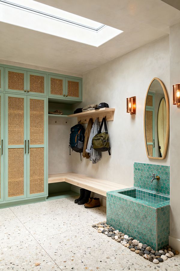 A bright mudroom illuminated by a large skylight features mint green cabinets with woven rattan fronts and brass knobs. A light wood L-shaped bench sits under a peg rail and shelf holding bags and hats. At right is an aqua scallop-tiled utility sink with a matching backsplash, edged by smooth beach pebbles. Off-white plaster walls, an oval wood-framed mirror, amber glass sconces, and a speckled terrazzo floor complete the coastal look, with a surfboard reflected in the mirror.