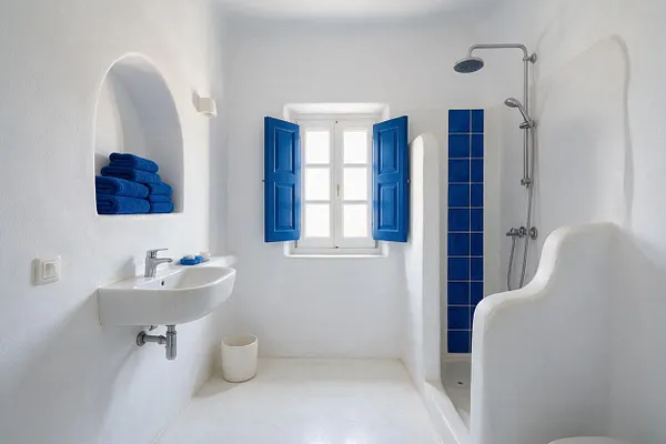 A bright Mediterranean-style bathroom with white stucco walls, a small wall-mounted sink, and a shower alcove framed by vivid blue tile. A window with matching blue shutters sits at the center, and built-in niche shelving holds folded blue towels.