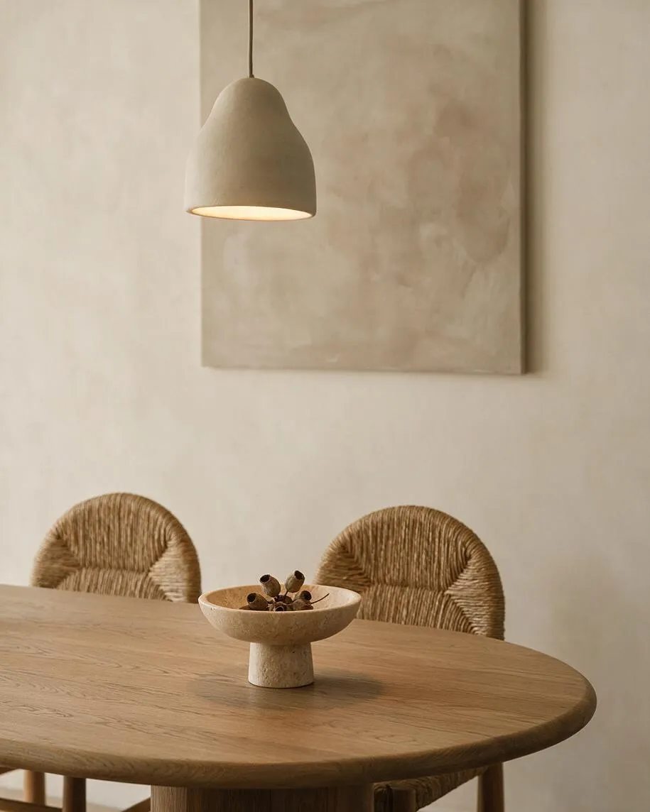 A serene dining nook with a round light oak table, two woven rope-back chairs, and a sculptural stone bowl centerpiece. Above, a soft beige pendant lamp hangs in front of a muted abstract wall canvas, all set against warm cream walls in a calm Organic Modern palette.