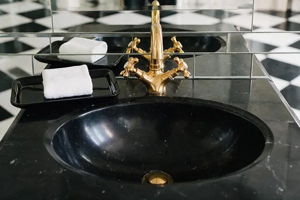 A bathroom vanity detail with a black marble countertop and integrated black sink, paired with a polished brass faucet. Behind it, mirrored wall tiles reflect the space, and a black-and-white checkerboard floor adds a bold graphic pattern. Two black trays with rolled white towels sit beside the sink.
