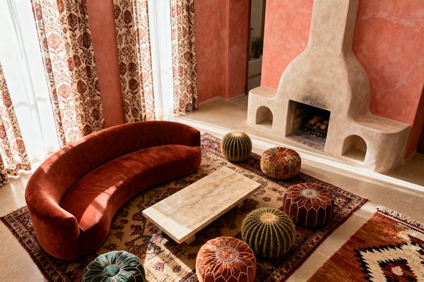 Sunlit living room with terracotta plaster walls and a sculptural adobe-style fireplace featuring arched niches and an open hearth. A curved rust velvet sofa surrounds a low travertine coffee table on layered patterned wool rugs; Moroccan leather poufs in rust, olive, and teal and two barrel cacti cluster near the table, while tall cream curtains with russet floral print filter light over a smooth beige floor.