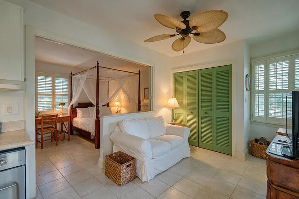 A tropical-style studio apartment with a white slipcovered loveseat, a canopy bed with sheer drapes in the sleeping area, and green louvered closet doors. The room features light tile floors, plantation shutters, woven baskets, wood furniture, and a ceiling fan with leaf-shaped blades.