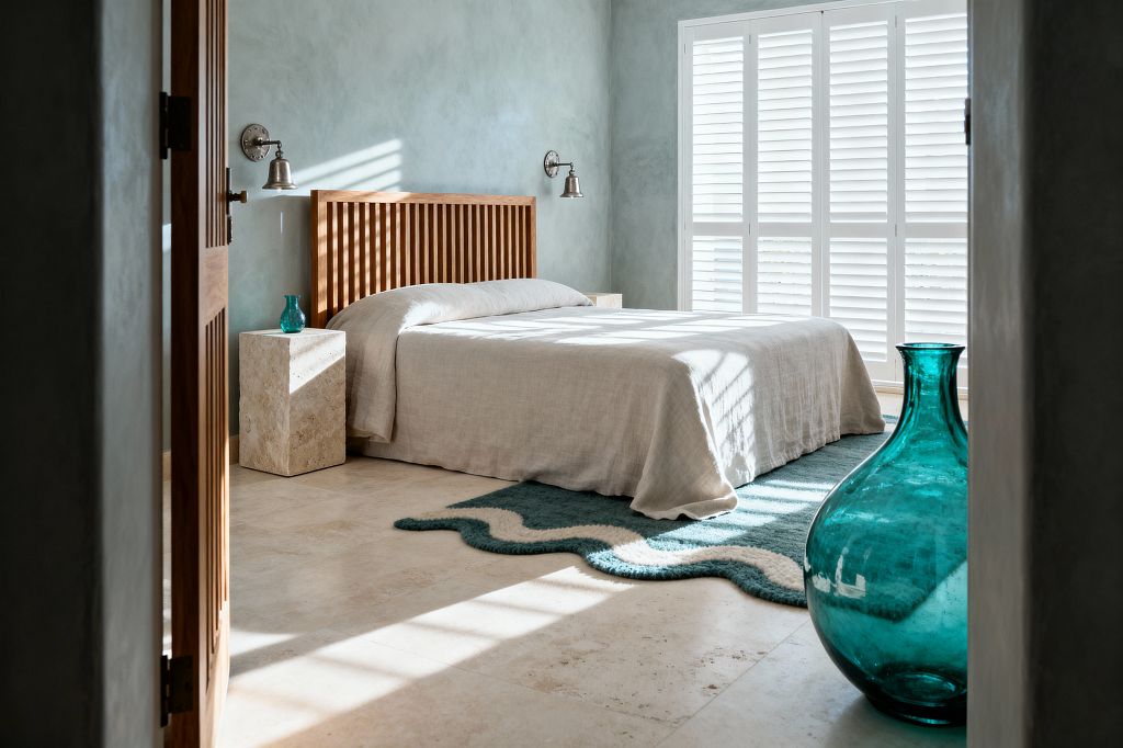 Sunlit coastal bedroom with a low bed dressed in natural linen against a slatted wood headboard. Travertine block nightstands flank the bed beneath brushed metal sconces, set on light stone flooring. Seafoam, limewashed walls and wide white plantation shutters fill the room with soft daylight and striped shadows. A teal wave-pattern wool rug sits under the bed, and a large teal blown-glass vase stands in the foreground.