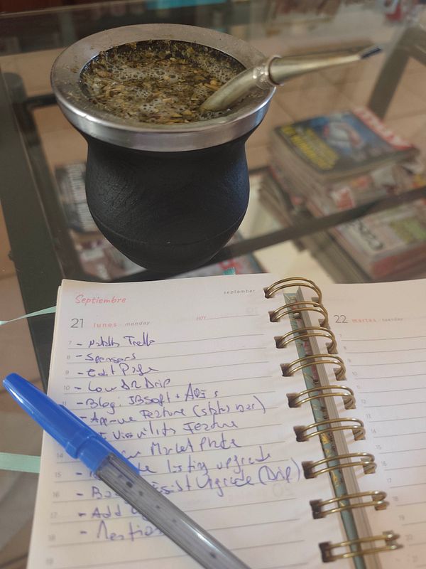 A black mate cup with a metal straw is placed on a glass table next to an open planner with handwritten to-do list for Monday, September 21, and a blue pen.