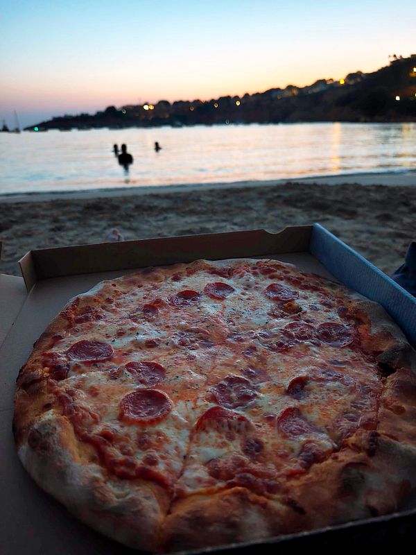 A person eating a pepperoni pizza slice at a sandy beach with waves and a cloudy sky in the background.