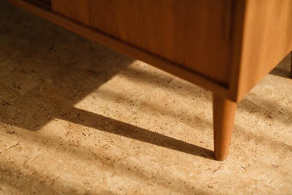 A close-up of a walnut mid-century modern cabinet with tapered legs casting long shadows onto a light travertine floor. The stone surface shows natural speckling and soft beige tones, creating a warm, retro-inspired interior detail.