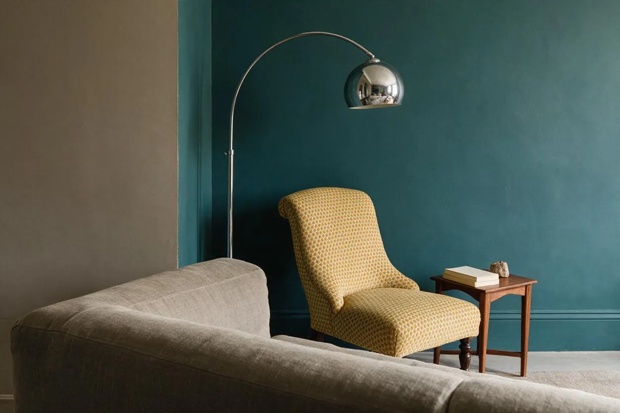 An eclectic living room vignette with a deep teal accent wall, a mustard patterned accent chair, and a chrome arched floor lamp. A neutral upholstered sofa fills the foreground, while a small wooden side table with books adds warmth beside the chair.