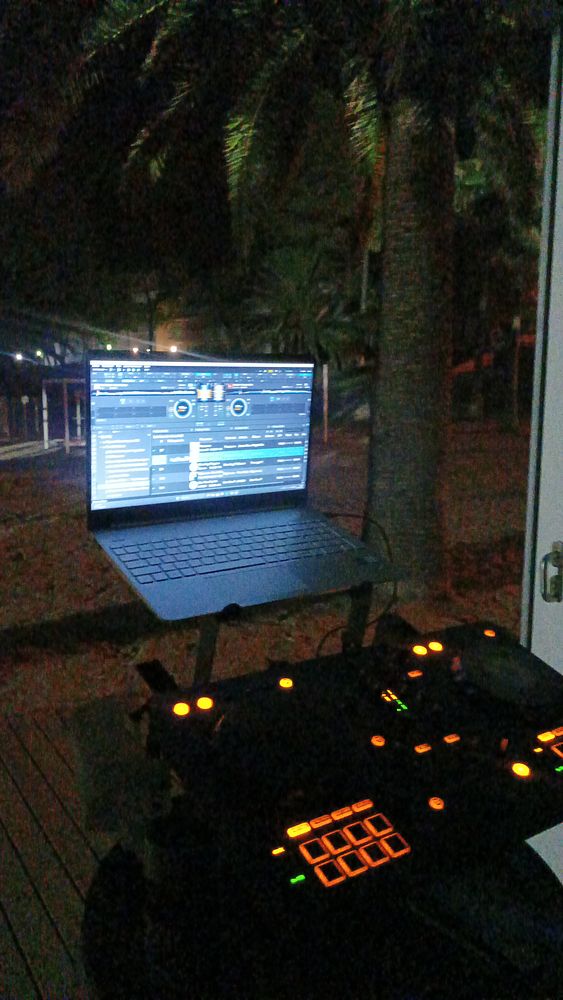 A DJ setup is illuminated at night, featuring a laptop and DJ controller surrounded by palm trees.
