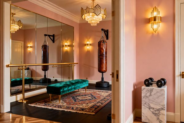 A blush-pink home gym features floor-to-ceiling mirrored panels with a polished brass ballet barre, an emerald tufted velvet bench, and a Persian-style rug layered over black rubber flooring. Art Deco crystal chandelier and matching brass sconces cast warm light on a cognac leather punching bag, while a white marble pedestal holds dumbbells near a doorway trimmed in cream molding.