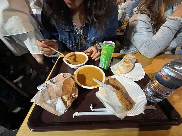 A person enjoys a meal at a soup festival, featuring soup, bread, and a drink.