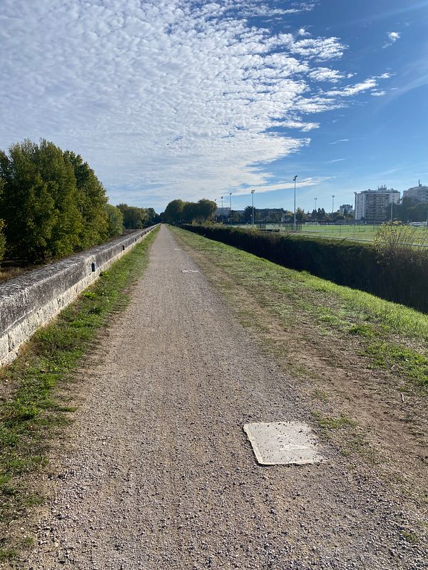 A straight gravel path flanked by greenery and a stone wall under a partly cloudy blue sky, ideal for walking.