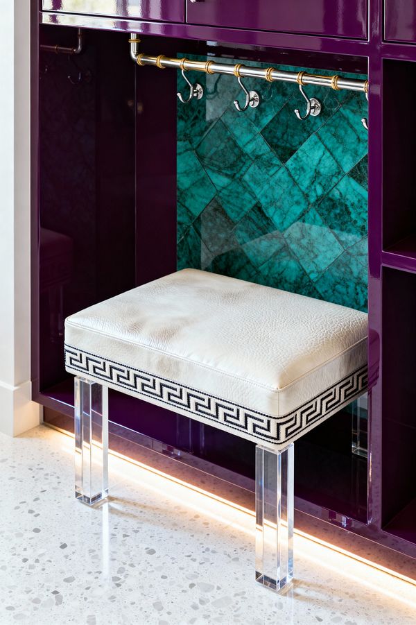 A mudroom nook features glossy plum lacquer cabinetry framing a bench with a white leather cushion edged in black Greek-key trim and supported by clear acrylic legs. Behind a brass-accented hanging rail with polished nickel hooks, emerald herringbone stone tiles create a jewel-toned backdrop, while warm LED toe-kick lighting highlights the light gray terrazzo floor with small aggregate flecks.