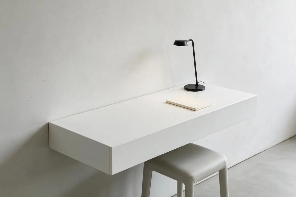 A minimalist workspace vignette shows a floating white wall-mounted desk against a plain white wall, with a small black task lamp and a slim notebook on top. A light gray stool tucks underneath, and the room is finished with a soft neutral palette and clean, modern lines.