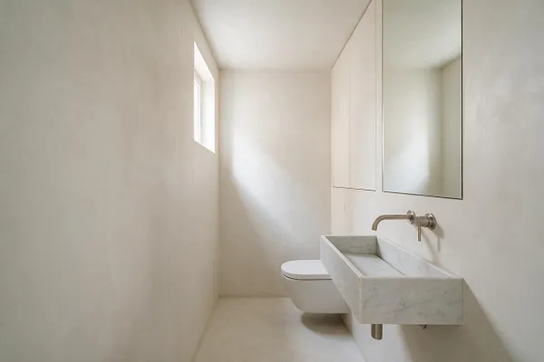 A narrow minimalist powder room with smooth off-white walls, a small square window, and a wall-mounted marble trough sink. A floating toilet sits beside the sink, and a large mirrored cabinet reflects soft natural light, creating a calm, spare atmosphere.