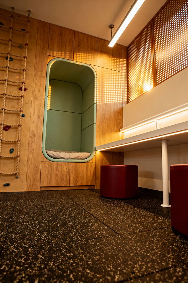 Modern children’s room with a built-in oak-veneer wall featuring a rounded, mint-green cushioned reading alcove above low drawers. To the left, a rope ladder and small climbing holds are mounted on the wooden panel. On the right, a long white desk with under-shelf LED lighting stands on a white metal leg, paired with two burgundy cylindrical stools. Perforated wood screens filter warm sunlight, and a black speckled rubber floor provides durable, safe footing.