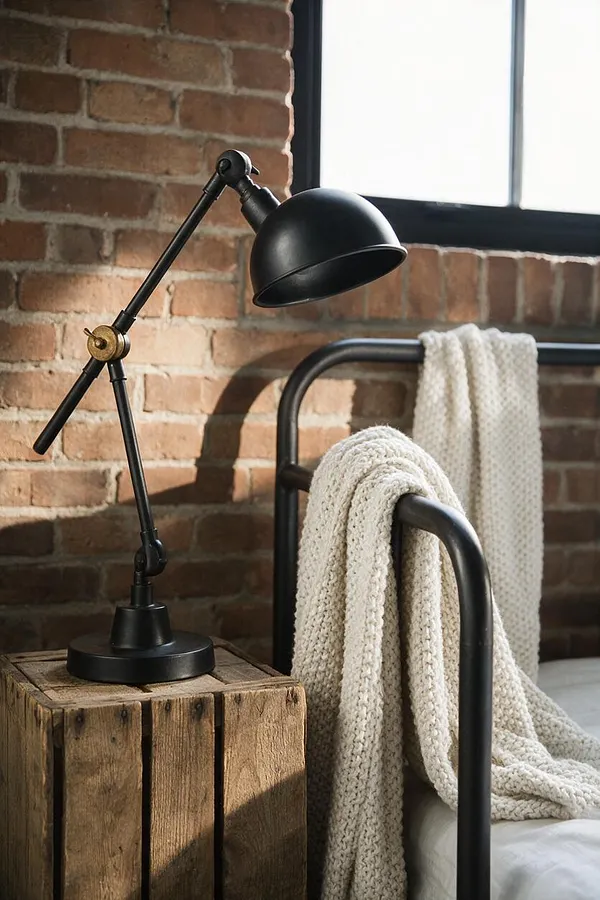 A cozy industrial bedroom vignette with an exposed brick wall, a black metal task lamp on a rustic wooden crate, and a dark metal bed frame. A chunky cream knit blanket is draped over the bed, and soft daylight enters through a black-framed window.
