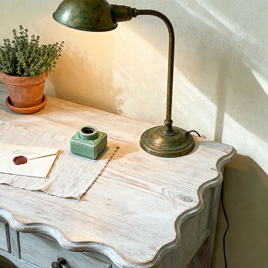 Close-up of a French Country home office vignette with a scalloped, whitewashed wood desk against a warm cream limewashed wall. An antique verdigris brass task lamp illuminates a frayed linen runner, a square green ceramic inkwell, and a cream envelope sealed with deep red wax. A small terracotta pot with a green herb sits at the corner. Soft daylight casts gentle shadows, highlighting worn edges and natural wood grain for a calm, timeworn atmosphere.